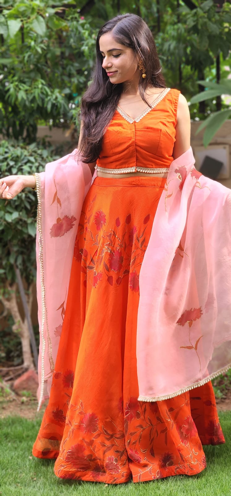 Woman in an orange traditional outfit with a pink dupatta standing outdoors.