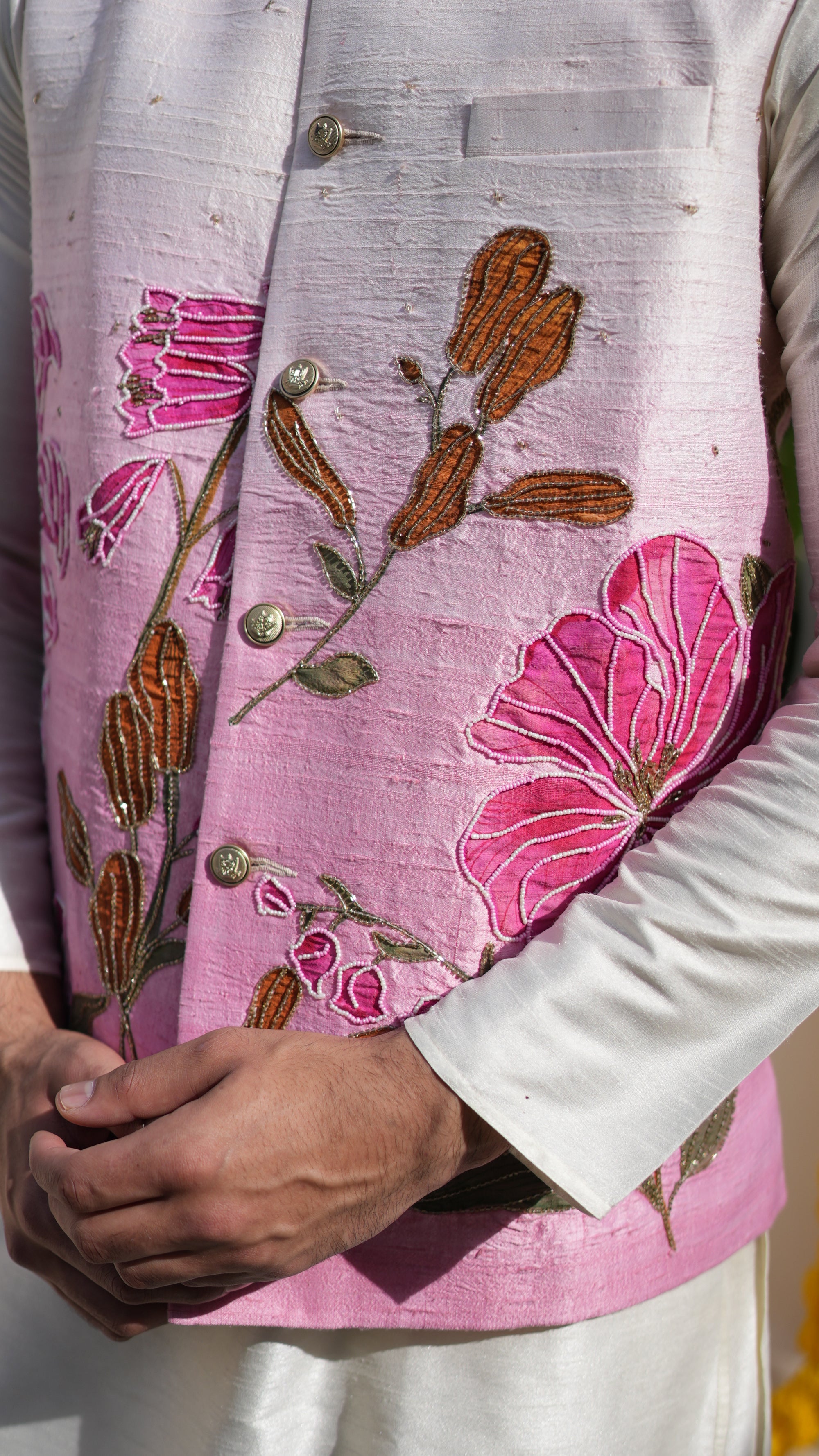 Person wearing a floral nehru jacket with a white kurta, standing in a haldi ceremony white background with green plants.