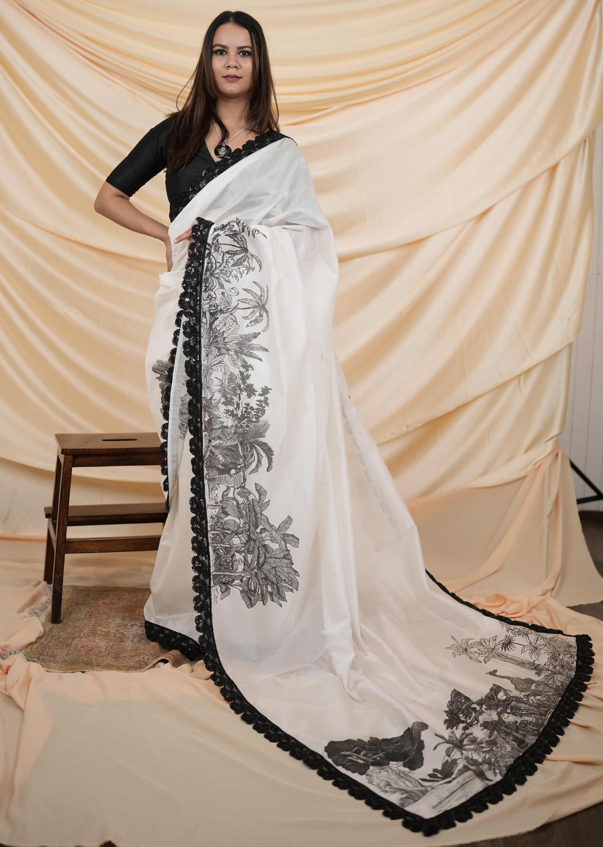 Woman wearing a white saree with black floral patterns against a beige curtain background