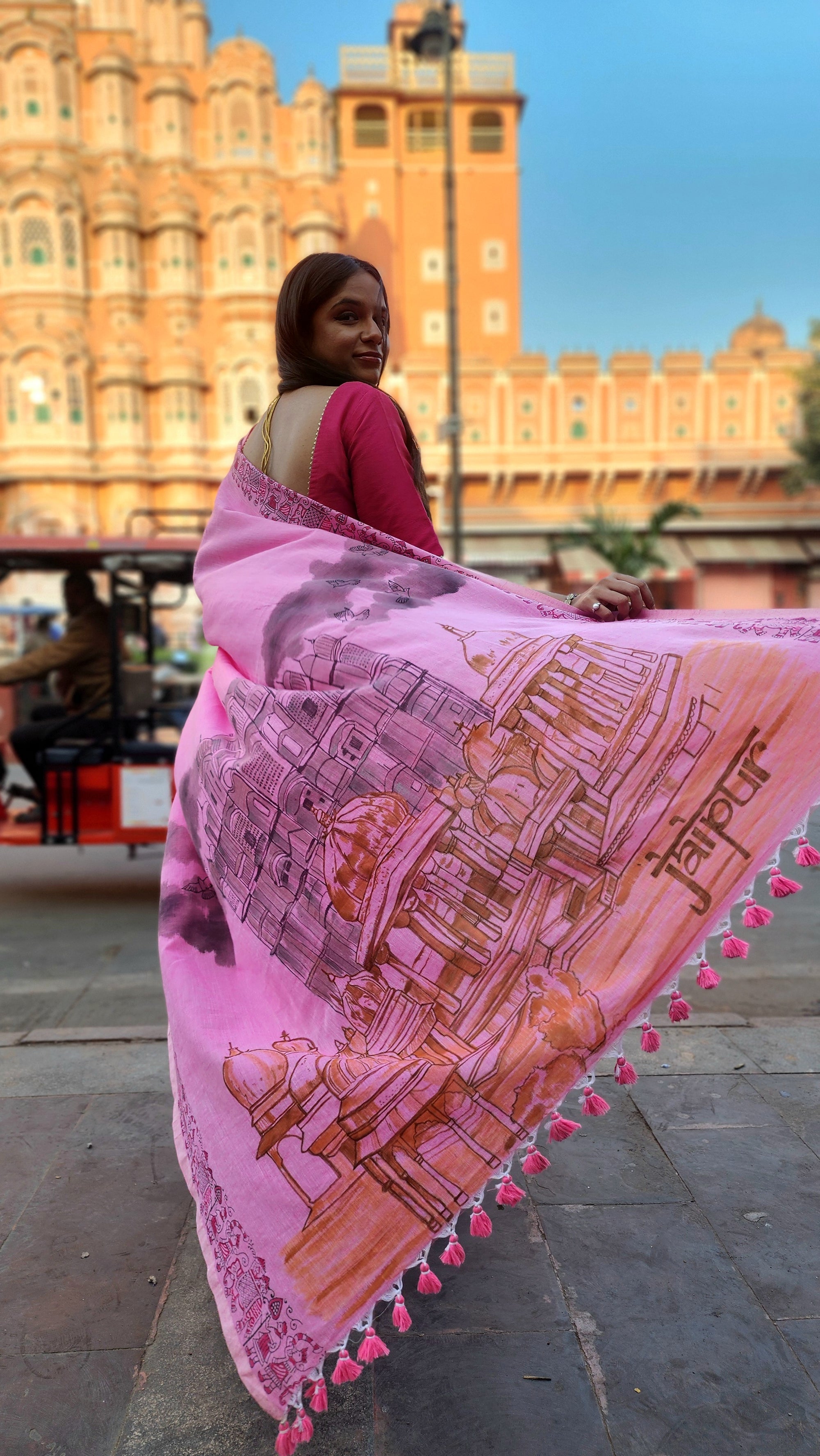 Woman holding a pink and orange patterned sari with a cityscape of Jaipur designed, standing in front of the Iconic Hawa Mahal.