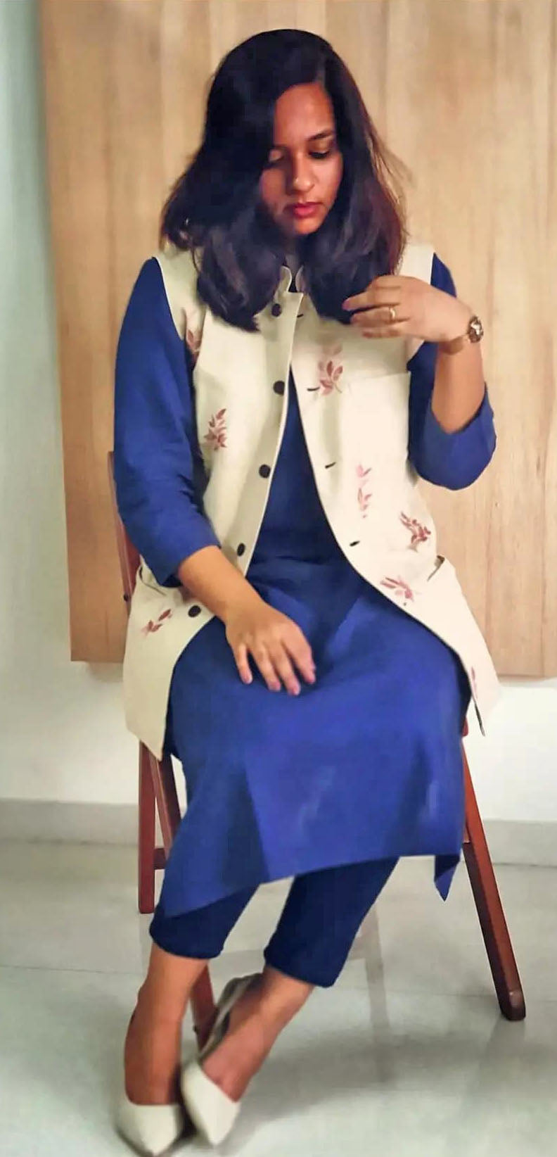 Woman wearing a blue kurta with a beige floral jacket sitting on a chair.