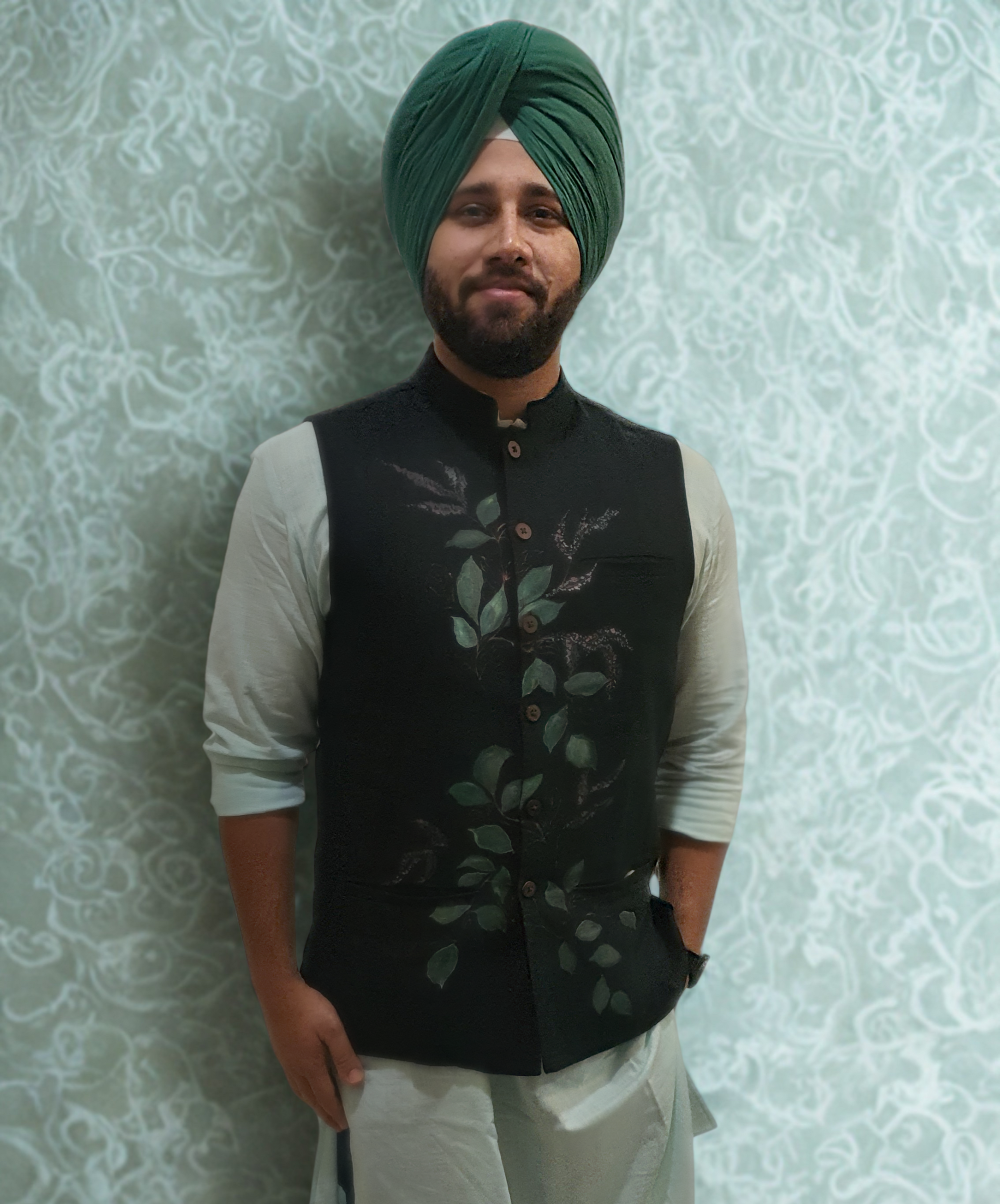 Man wearing a black hand painted nehru jacket  over a light green kurta with a green turban against a light patterned wall.