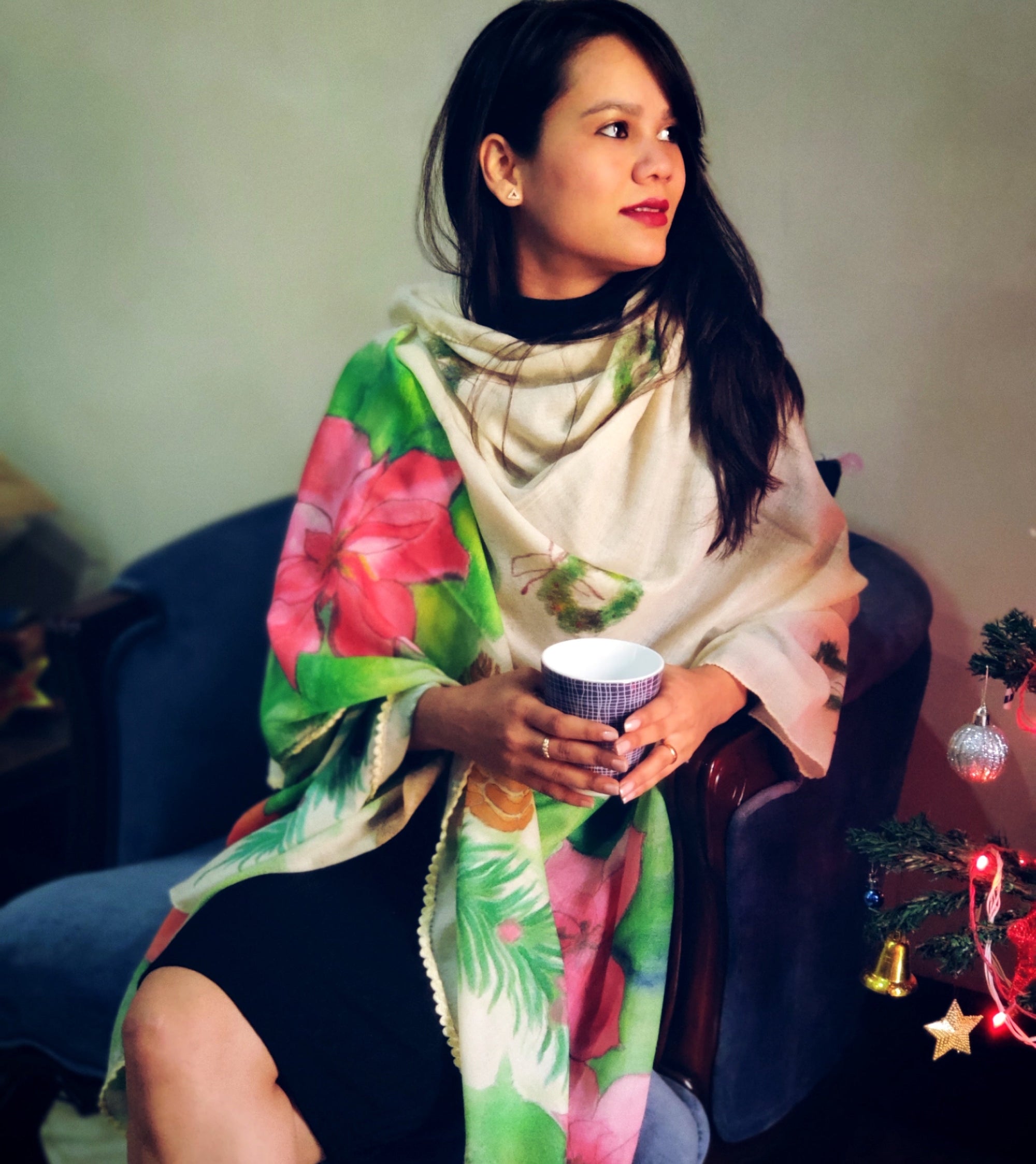 Woman wearing a hand painted floral scarf holding a cup, sitting on a couch with Christmas decorations.