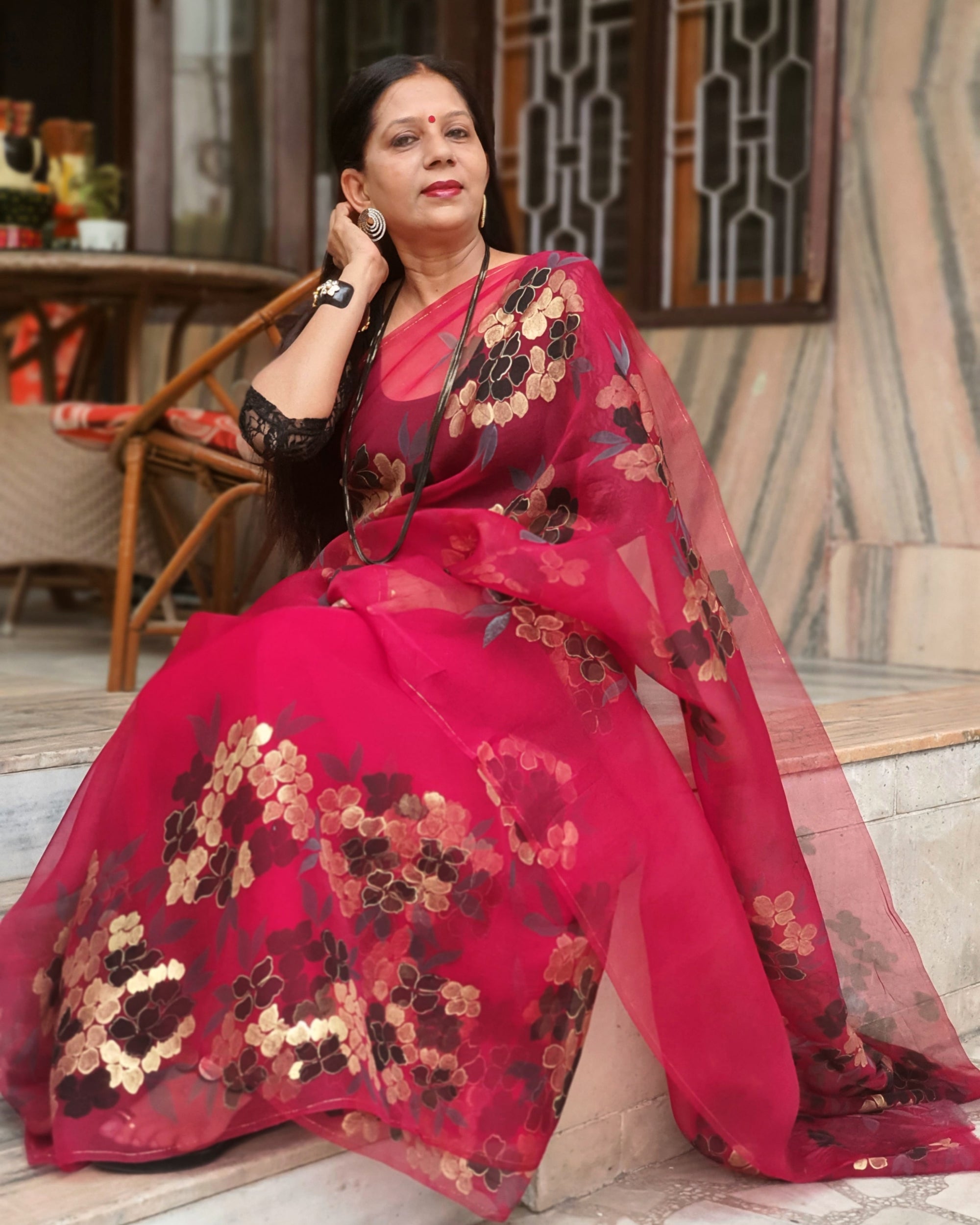 Woman wearing a red saree with floral patterns sitting outdoors.
