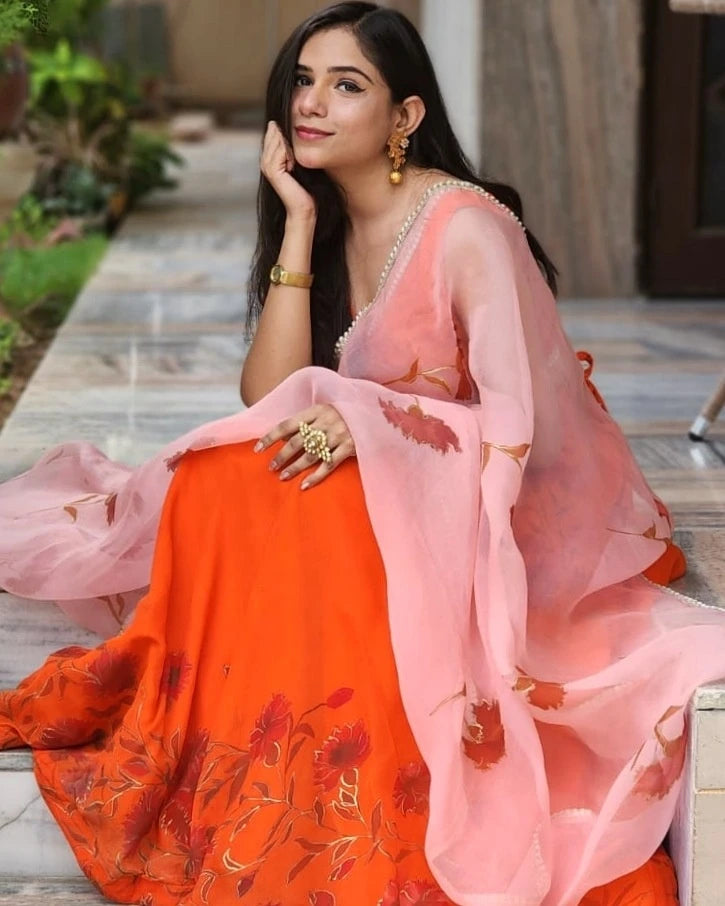 Woman wearing a traditional orange and pink saree with floral patterns, sitting outdoors.