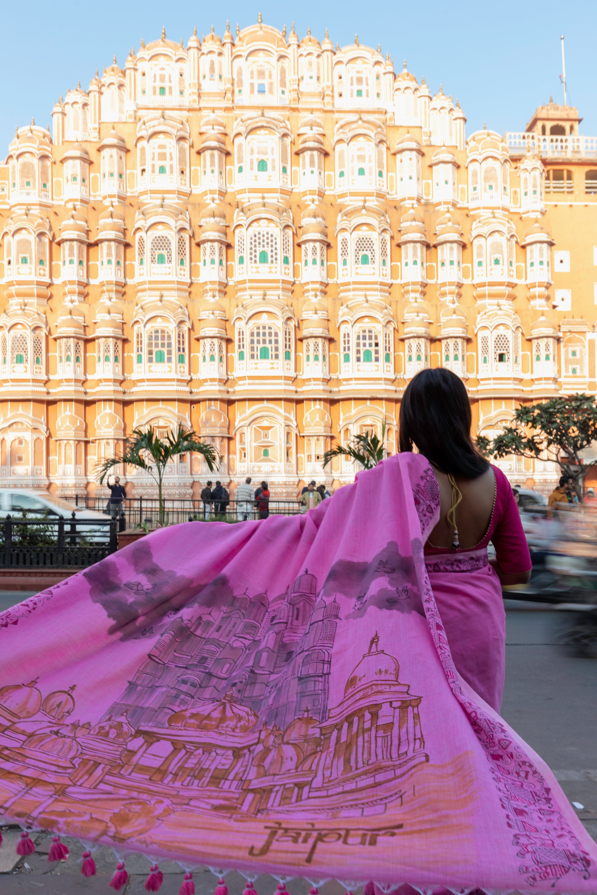 Woman in a pink saree with a detailed design walking in front of the Hawa Mahal in Jaipur, India.