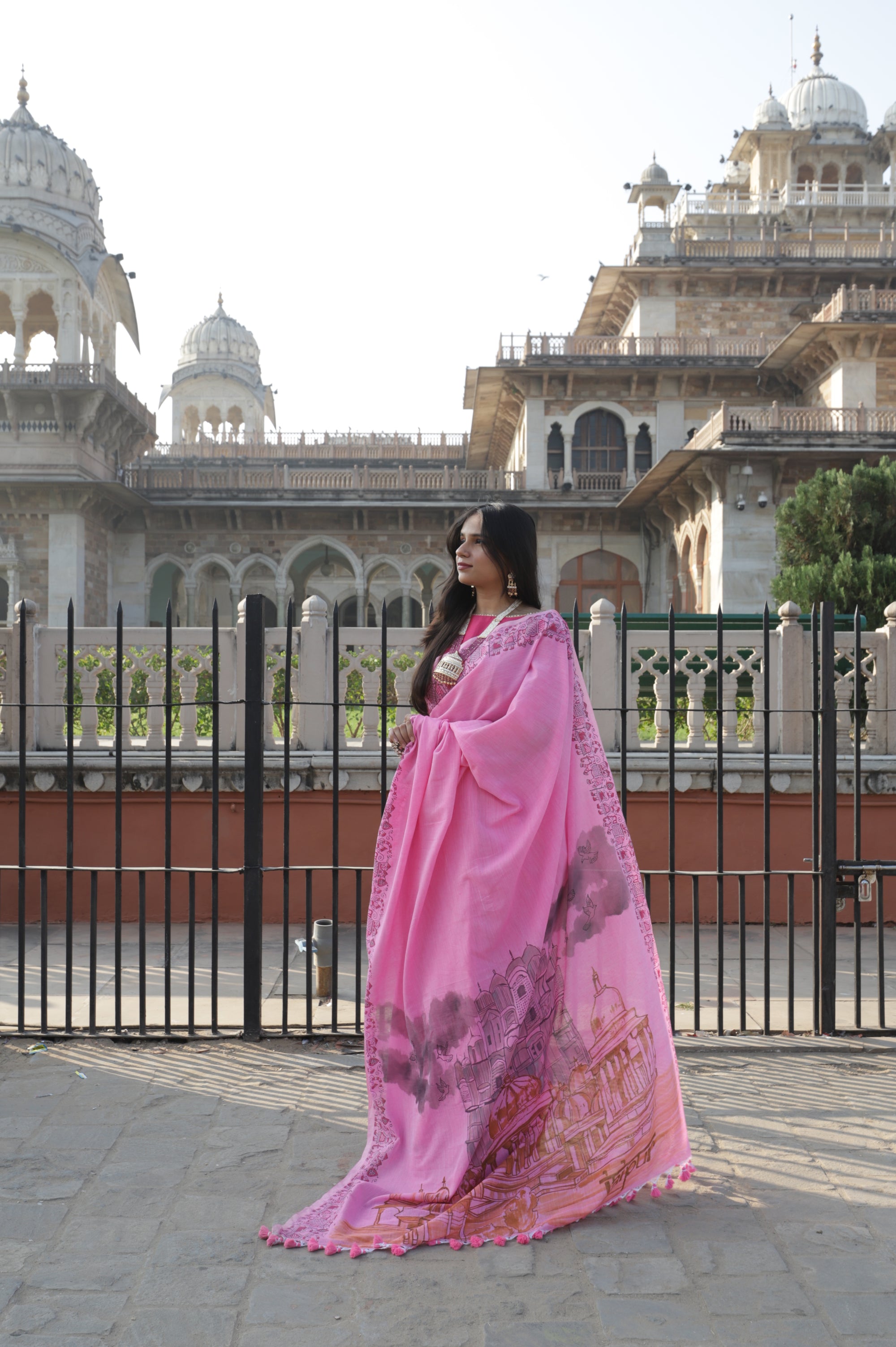 A woman wearing a pink sari with the iconic Hawa Mahal painted in front of a large Alber Hall building in Jaipur.
