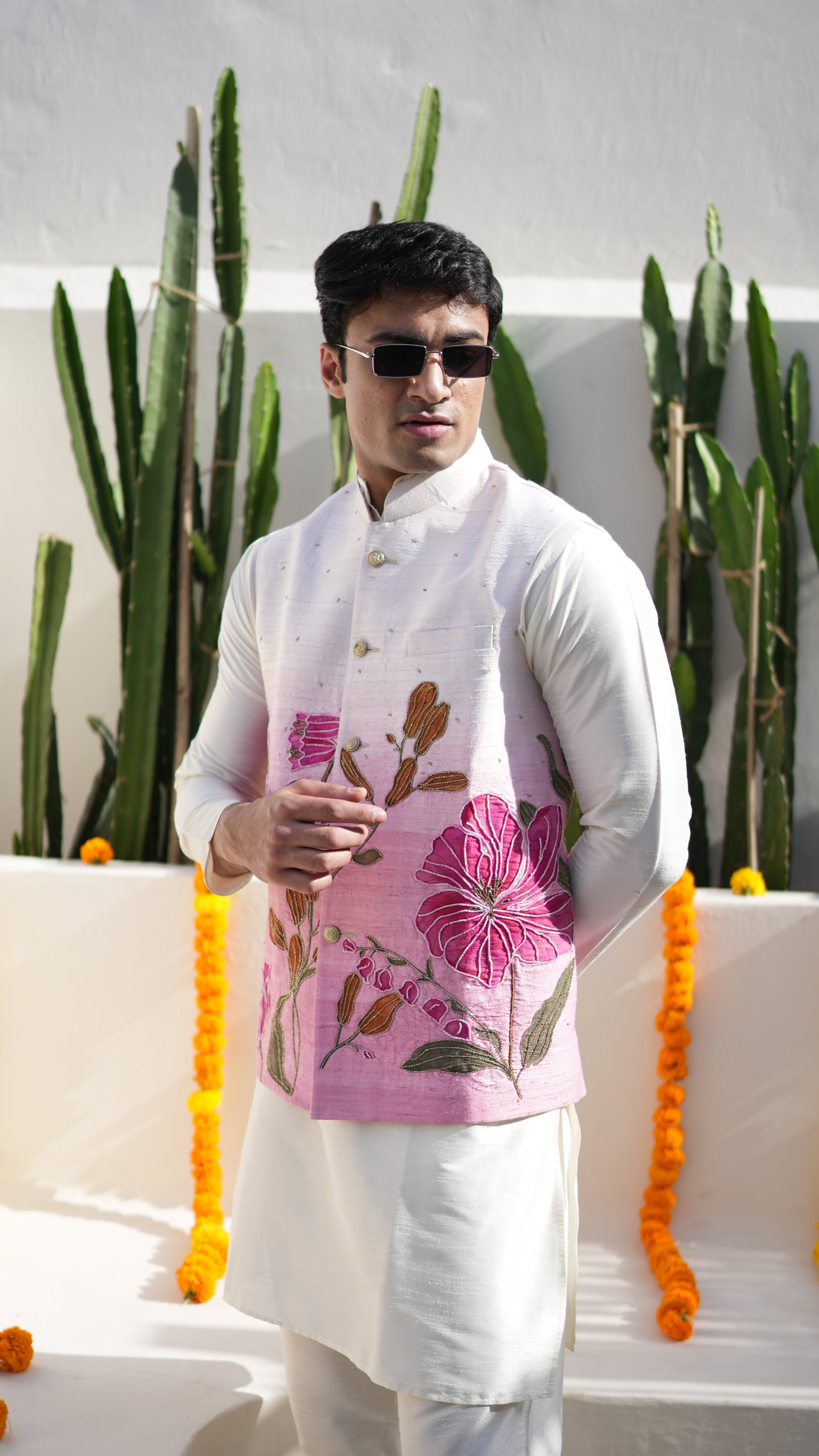 Person wearing a floral nehru jacket with a white kurta, standing in a haldi ceremony white background with green plants.