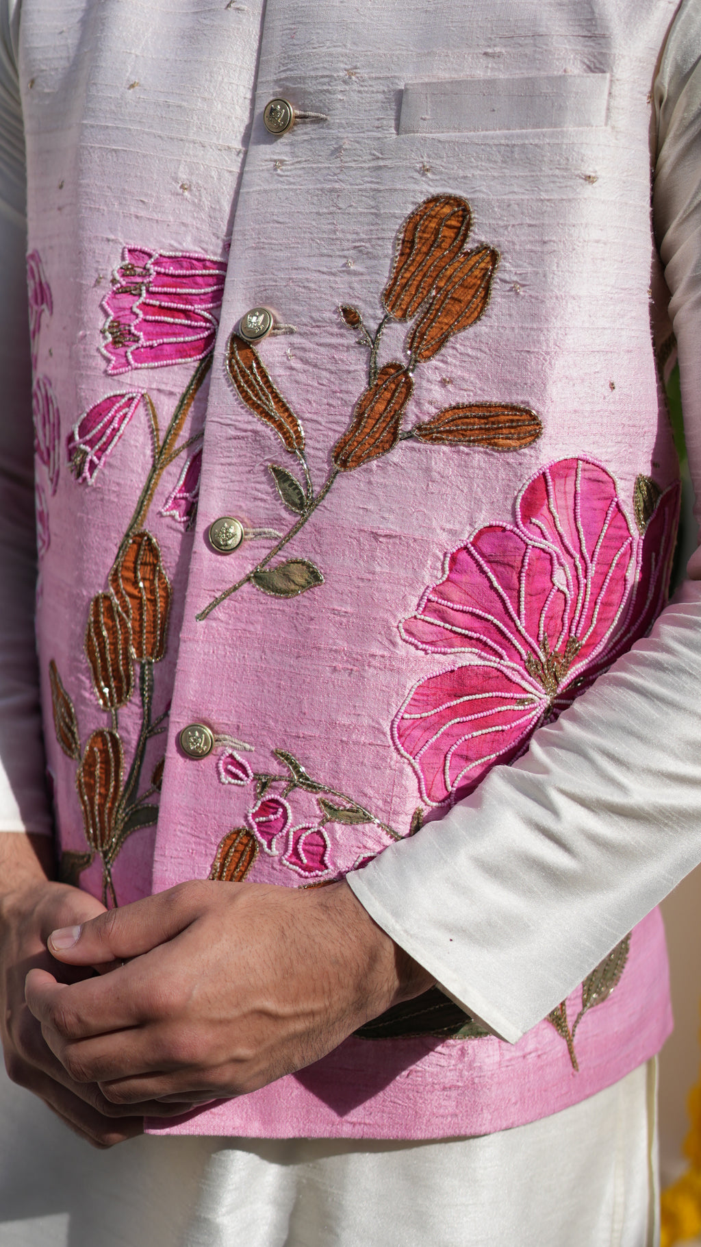 Person wearing a floral nehru jacket with a white kurta, standing in a haldi ceremony white background with green plants.