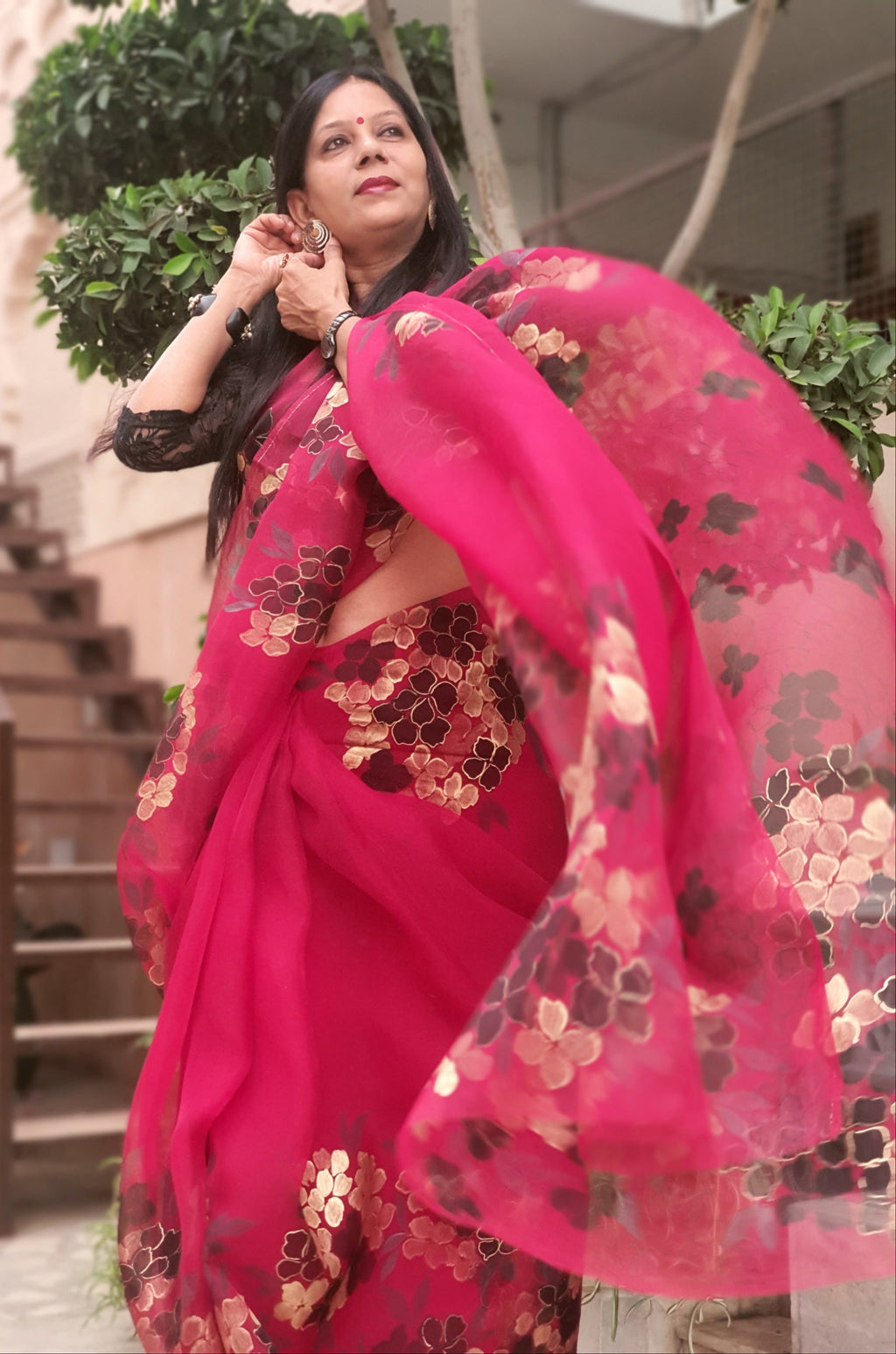 Woman in a bright pink saree with floral patterns, standing outdoors.
