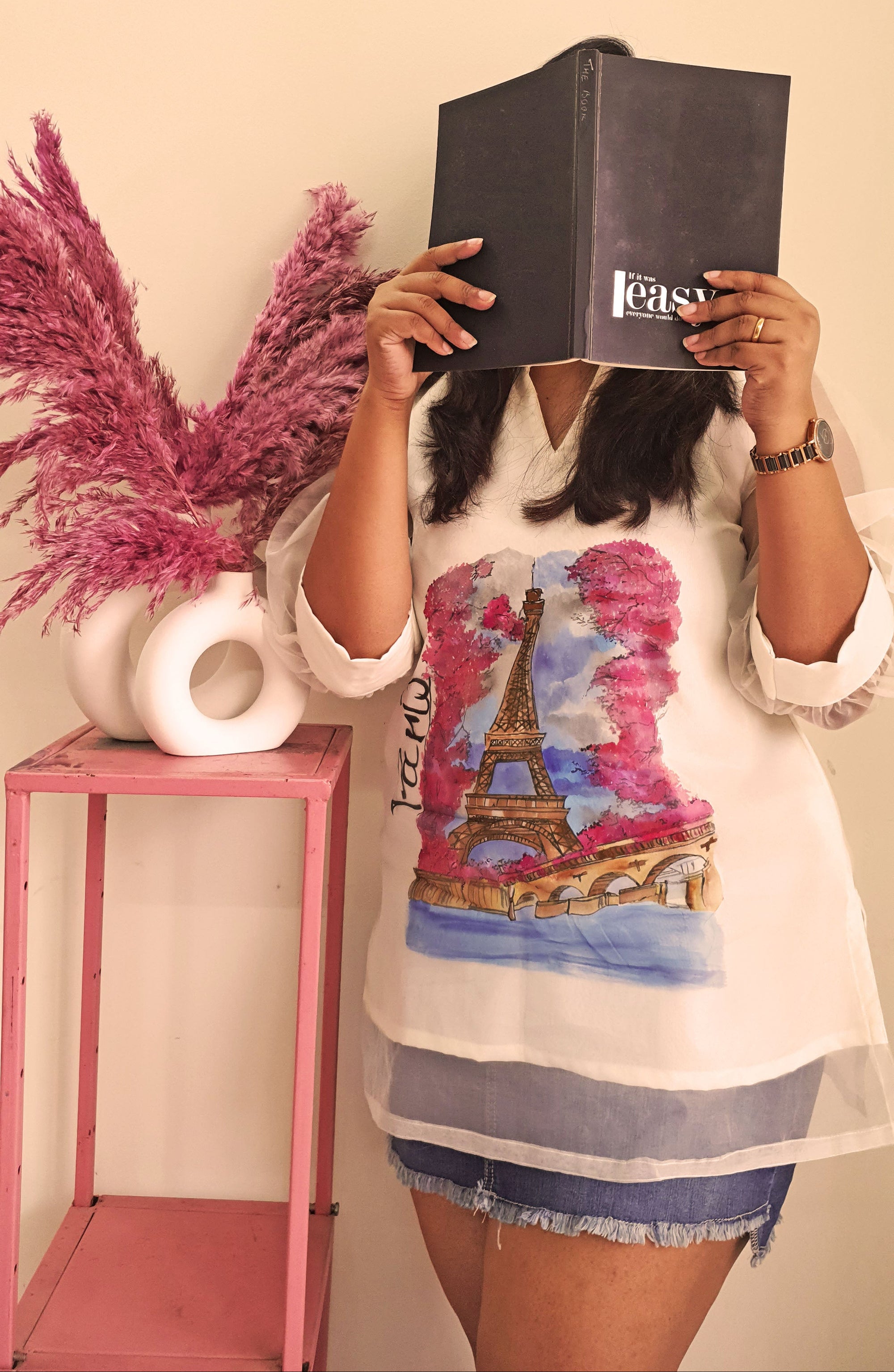 Person wearing a SHIRT with a Paris design, holding a book over their face, standing next to a pink table with a vase of pink flowers.