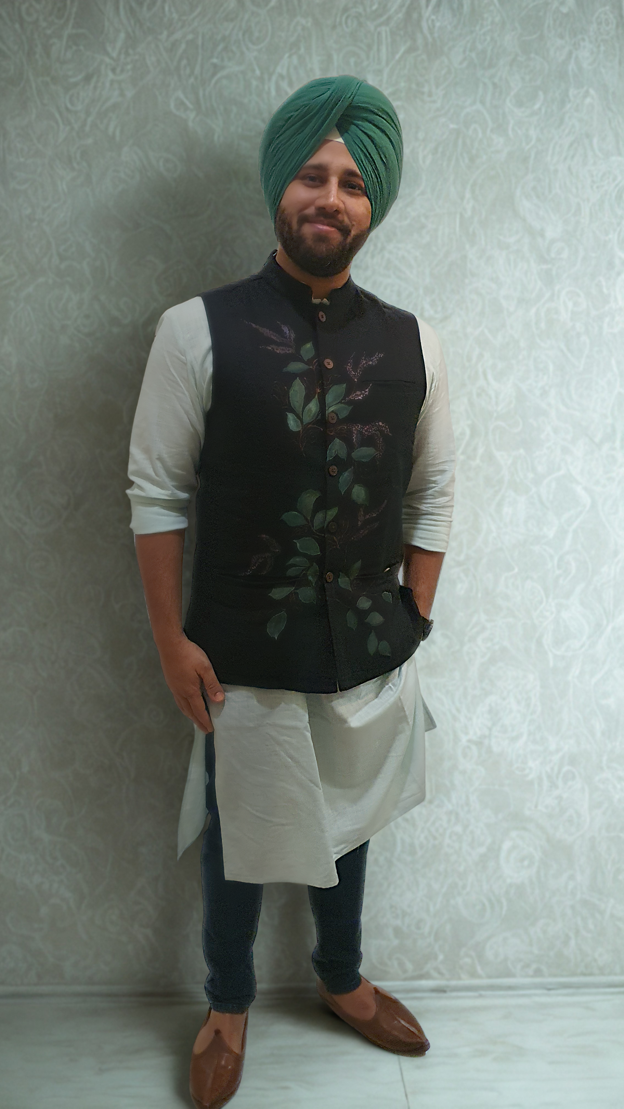 Man wearing a black hand painted nehru jacket  over a light green kurta with a green turban against a light patterned wall